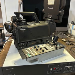 A 6 Panasonic AK-UC4000GSJ 4.4K Studio Camera System USED setup, with camera and monitor atop a Panasonic control unit, showcases quality preowned broadcast gear on a cluttered desk with a computer in the background.