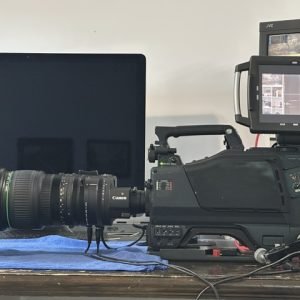 Professional video camera with an external monitor mounted on a desk, lenses facing left on a blue towel on the table.